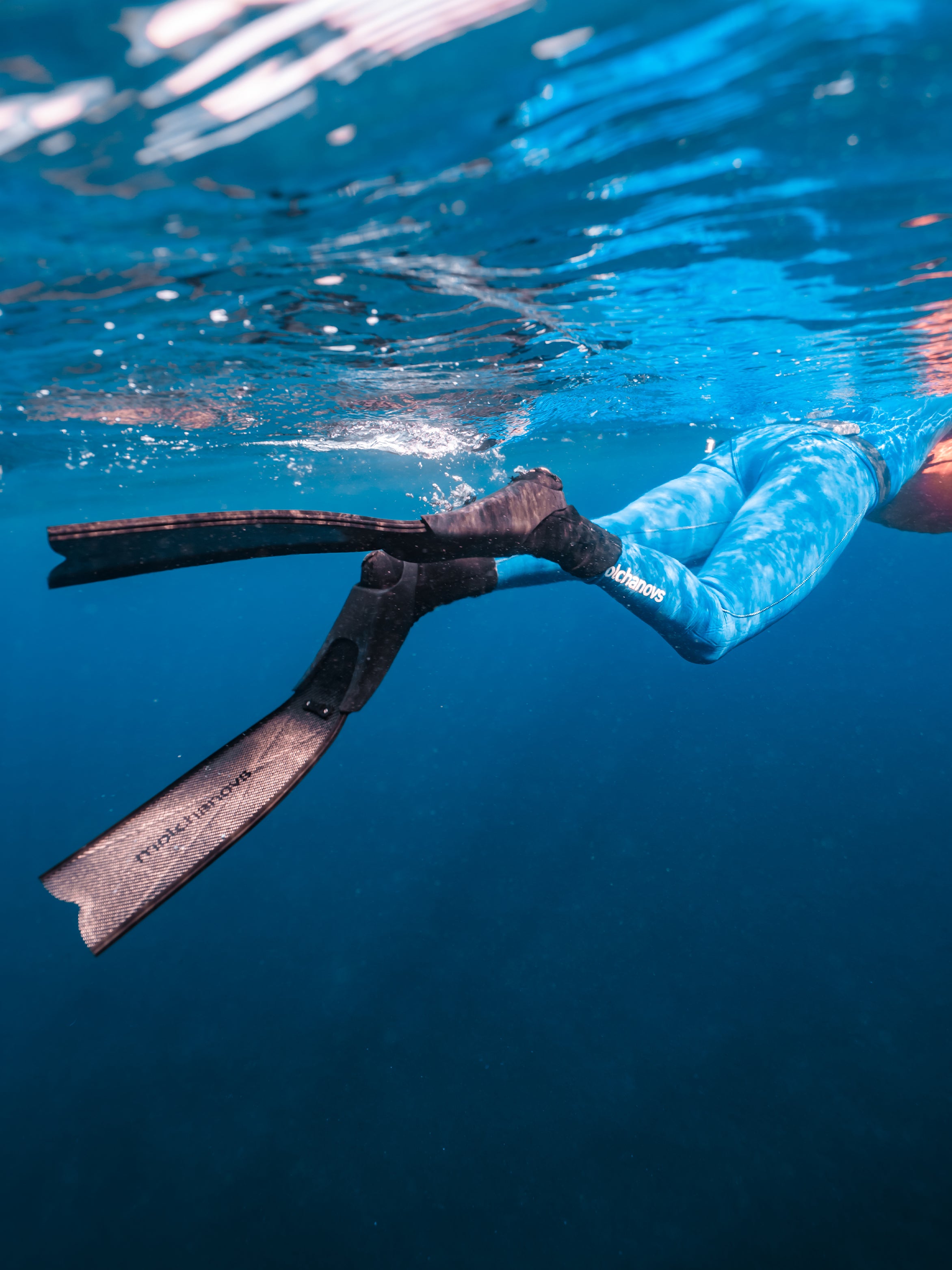 Underwater shot of a freediver kicking with Molchanovs short carbon bifins, legs mid-stroke near the surface in open water