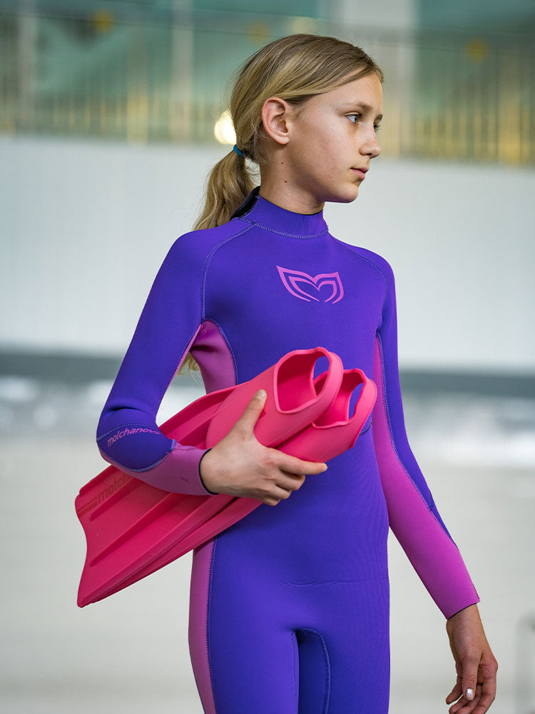 Girl in a purple and pink Molchanovs 2.5mm kids freediving wetsuit holding pink Molchanovs fins at an indoor pool