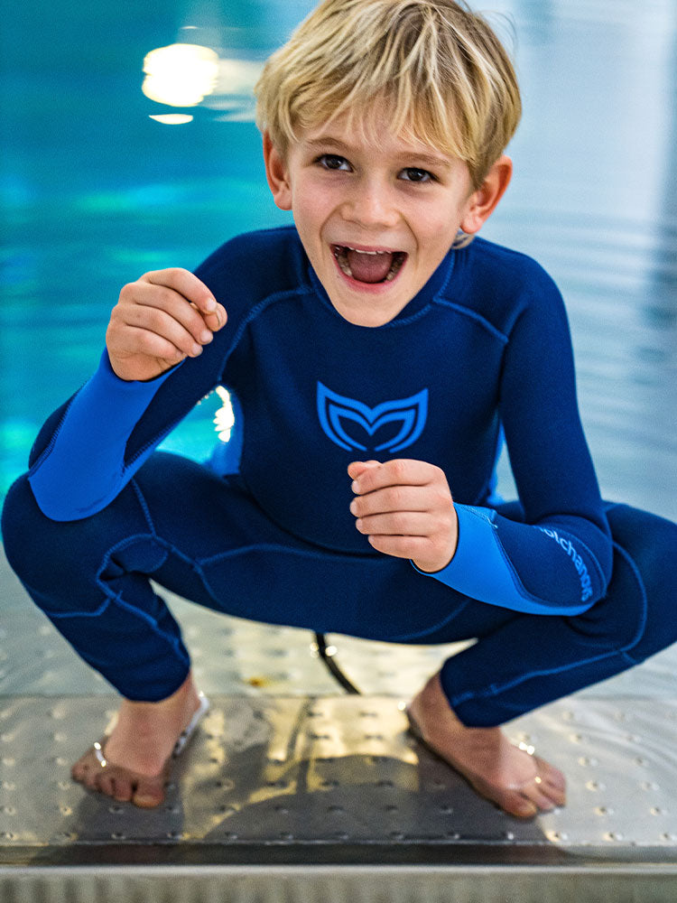 Young boy crouching at the pool edge wearing the Molchanovs 2.5mm kids wetsuit in navy blue, smiling