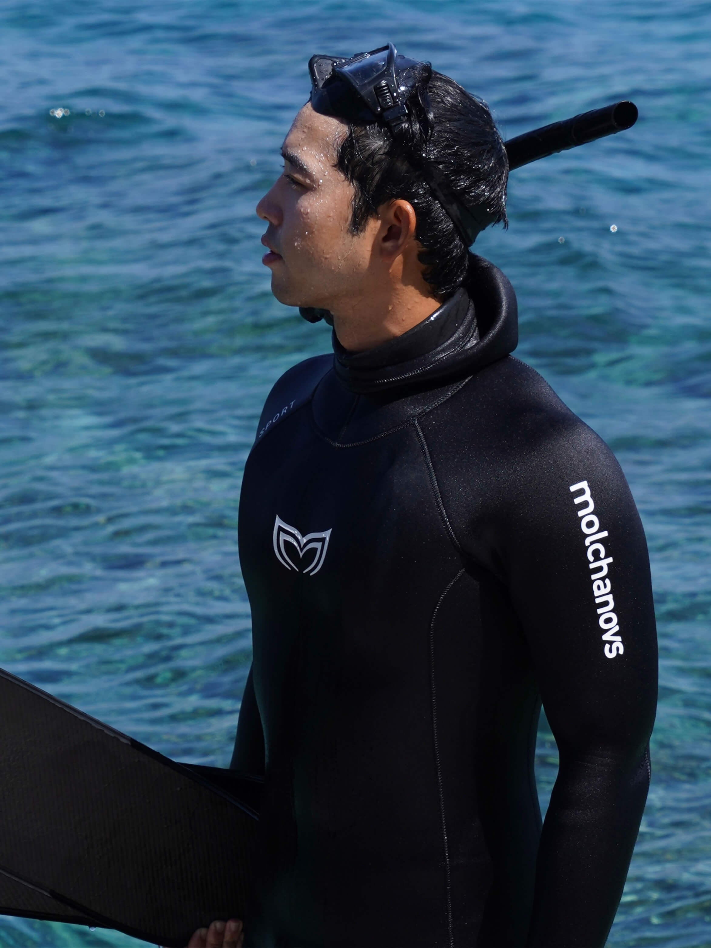 Male freediver in a black Molchanovs SPORT double-lined wetsuit holding fins at the water's edge, mask and snorkel on head