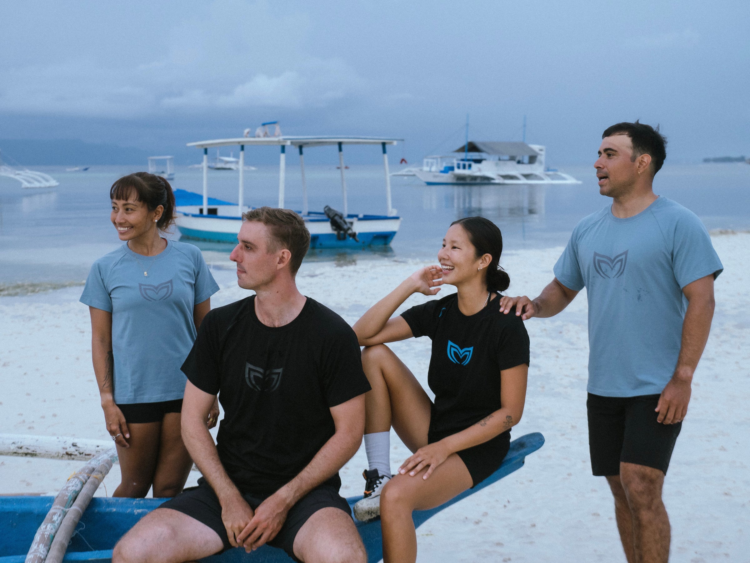 Group of four athletes wearing Molchanovs branded t-shirts on a tropical beach with outrigger boats in the background