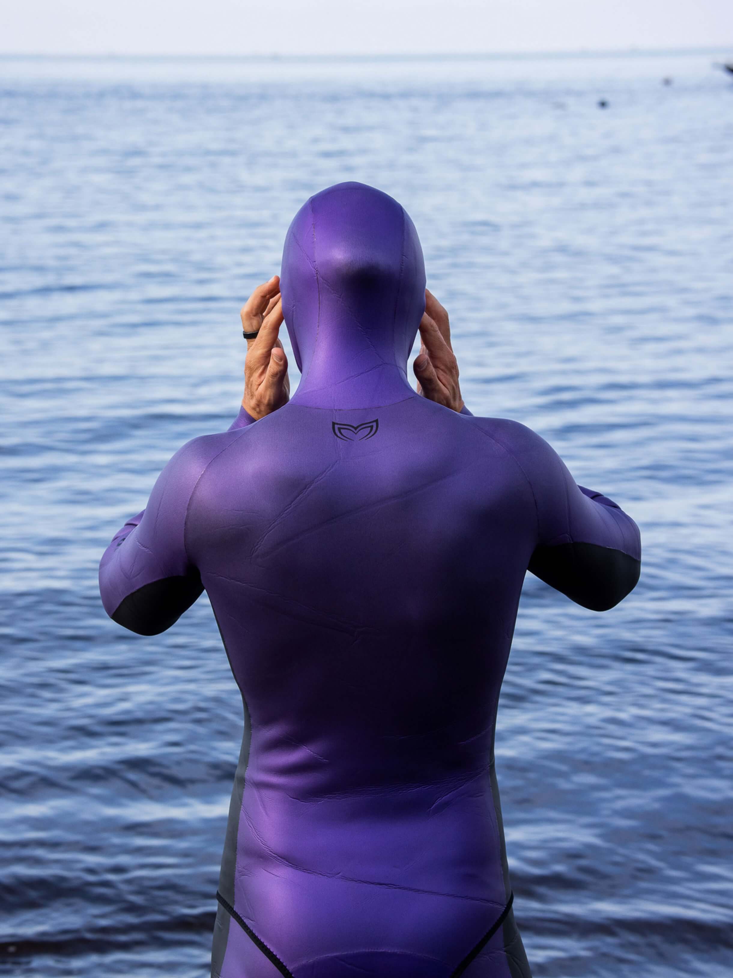 Freediver pulling on the hood of the Molchanovs Natalia's Legacy purple SPORT wetsuit 1.5mm, rear view at the coast