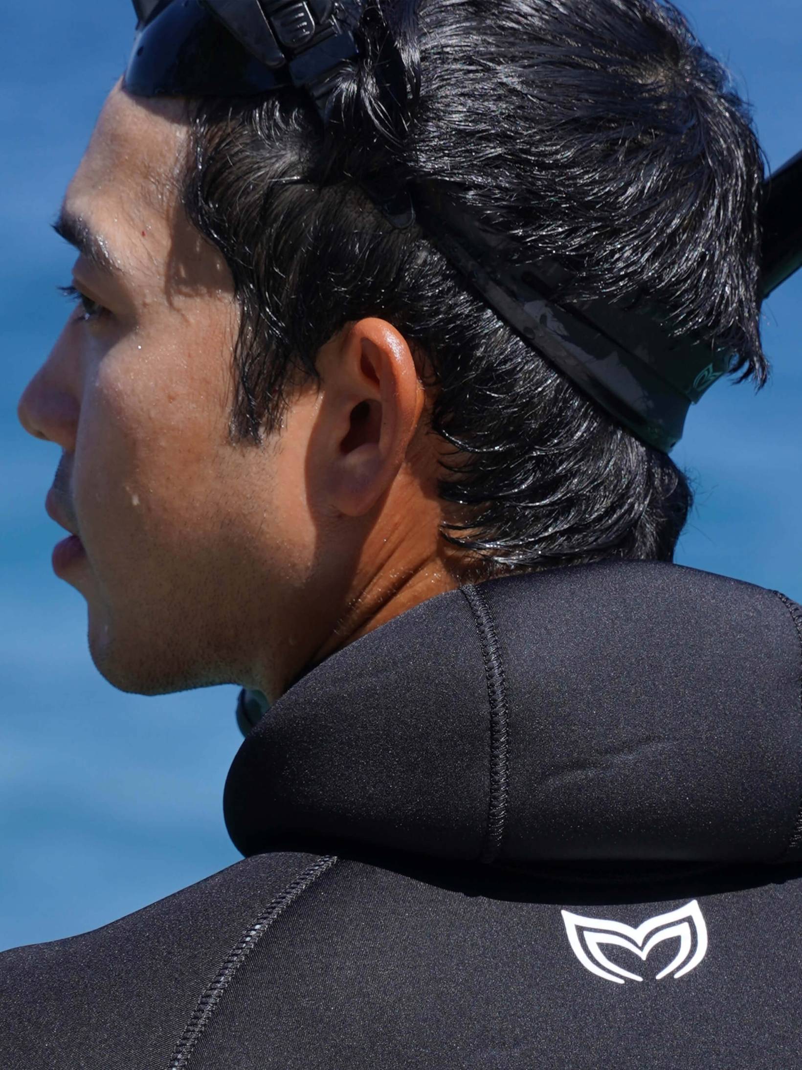 Male freediver in a black Molchanovs SPORT double-lined wetsuit, hood down, showing the hooded collar and shoulder seams from behind