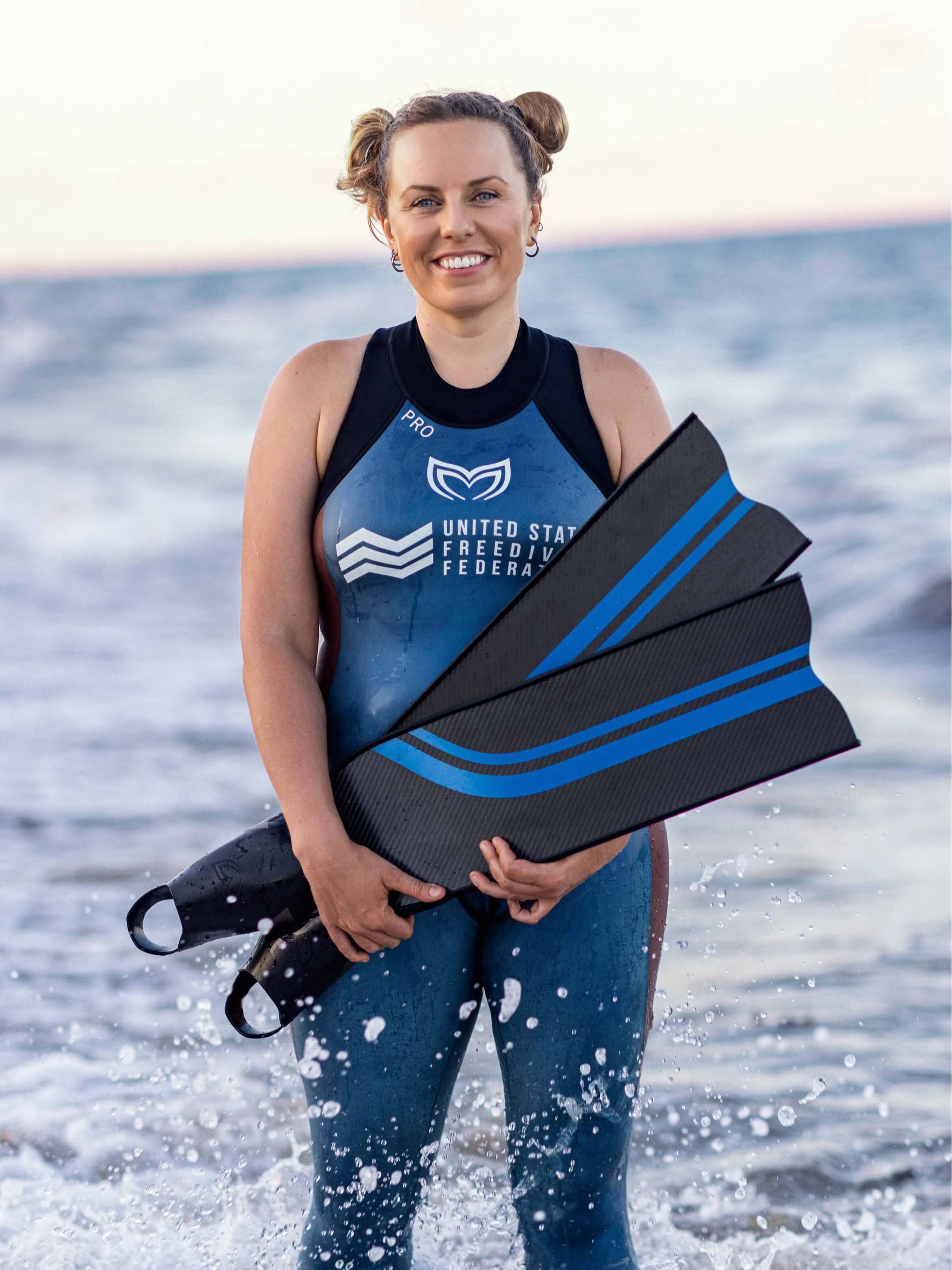Female freediver wearing a blue Molchanovs PRO women's wetsuit and United States Freediving Federation logo, holding carbon bifins at the shoreline