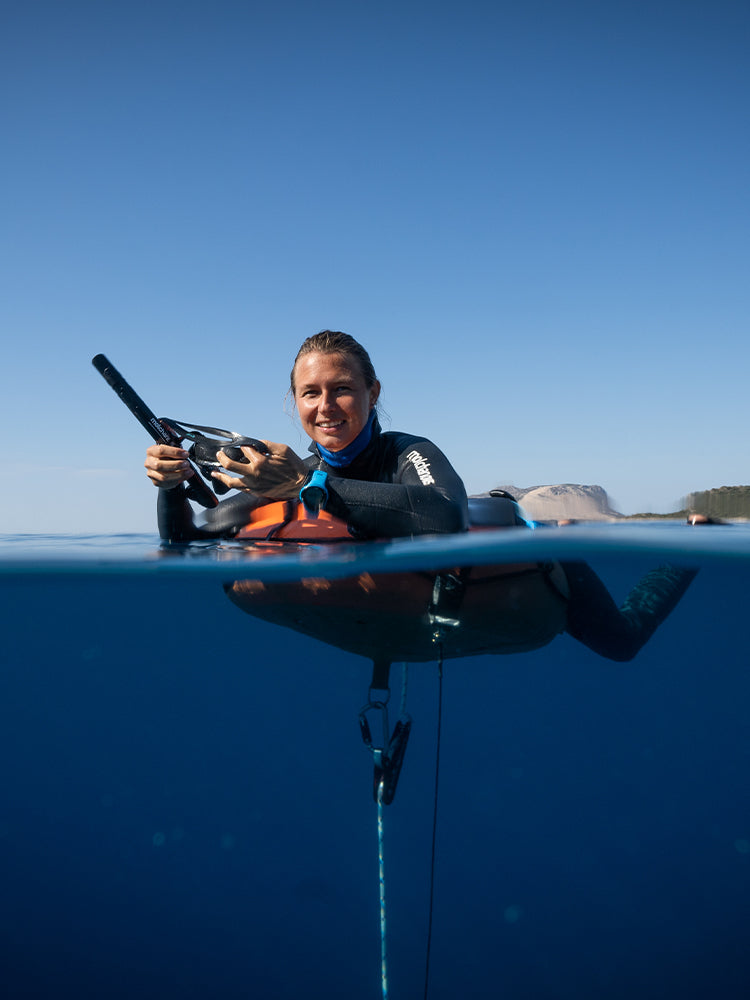 Freediver in a Molchanovs wetsuit resting on a PRO freediving buoy at the surface in open water, split above-below shot