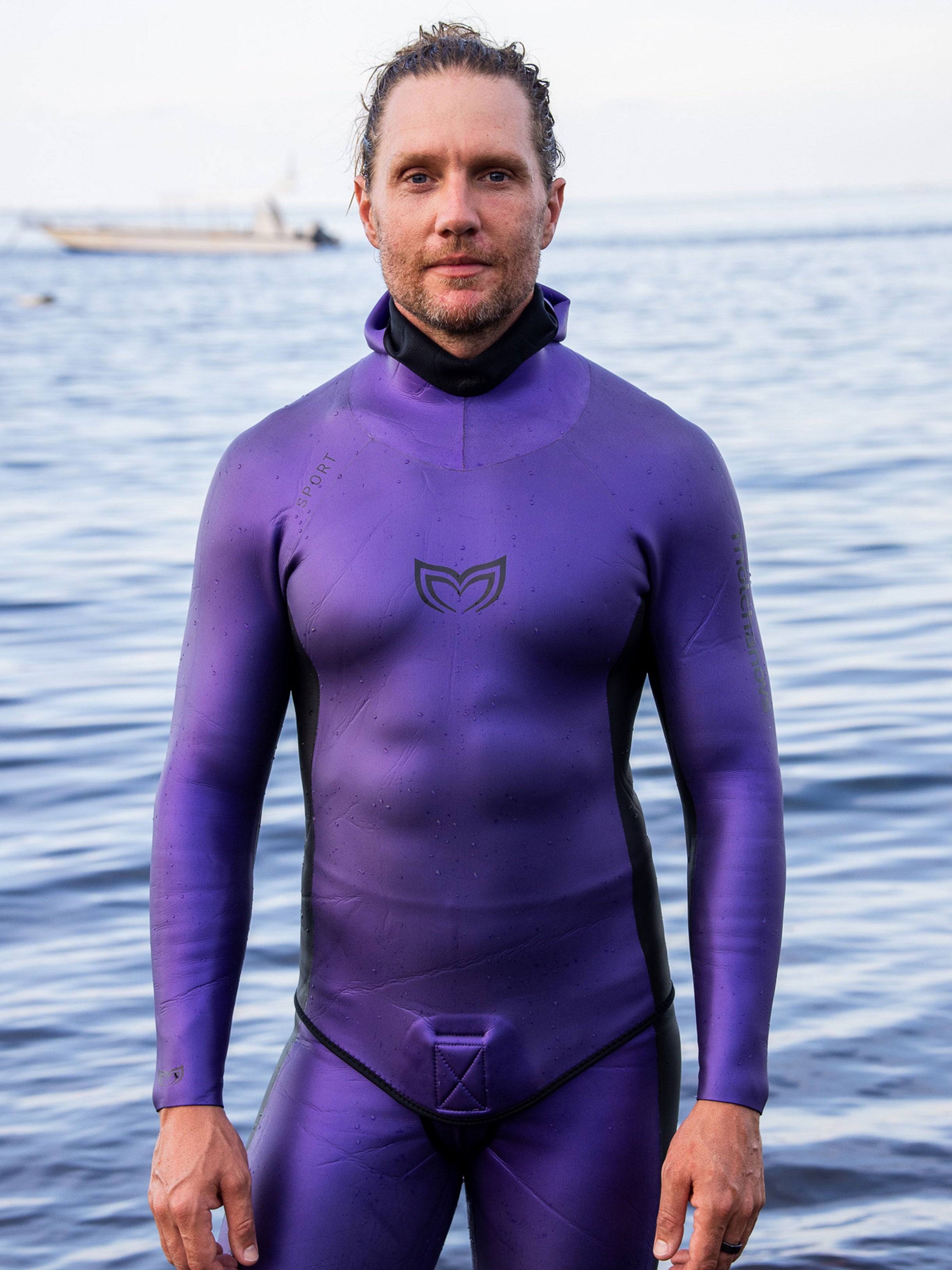 Male freediver wearing the Molchanovs SPORT purple smooth skin wetsuit with hood, standing at the water's edge