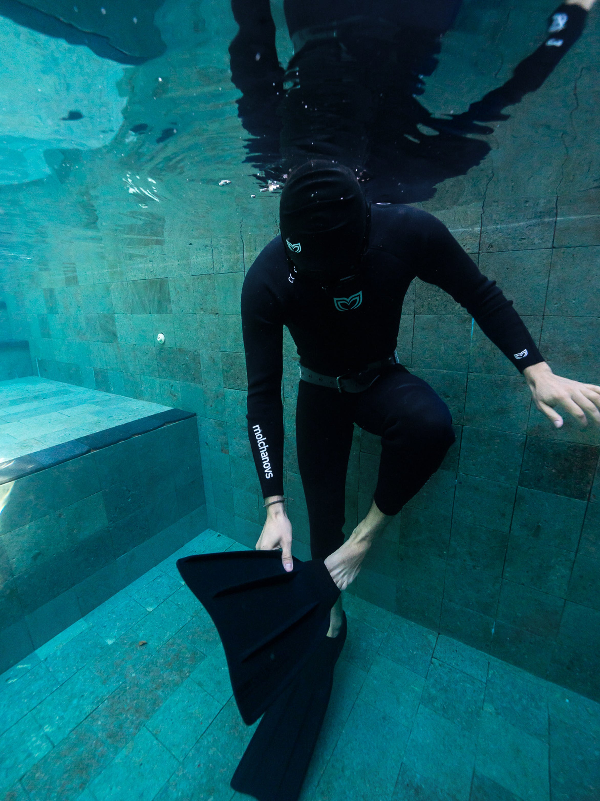 Freediver in Molchanovs CORE one piece wetsuit adjusting a monofin underwater in a pool