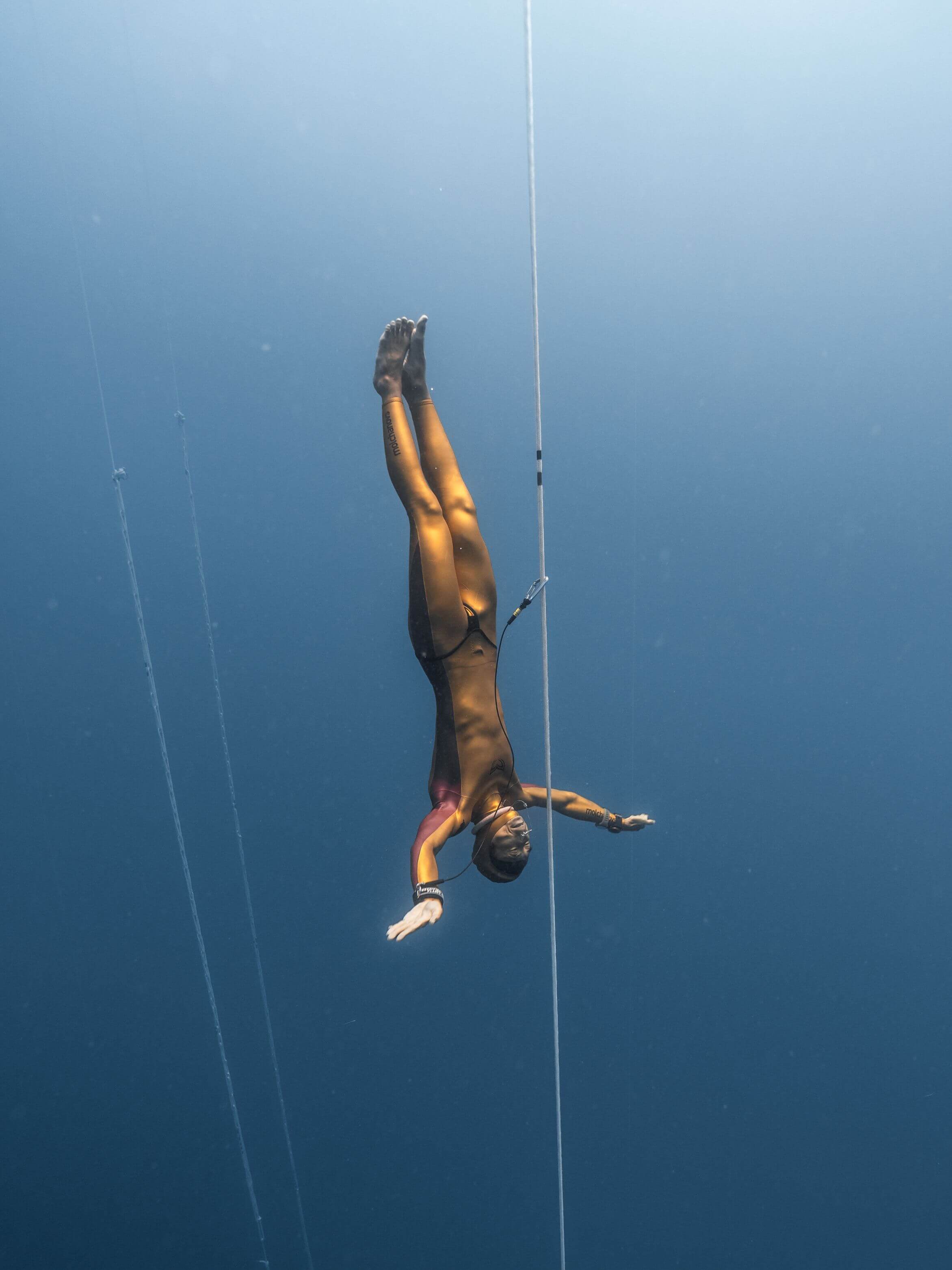 Freediver in a gold Molchanovs PRO wetsuit gliding back up along a competition line in deep blue water